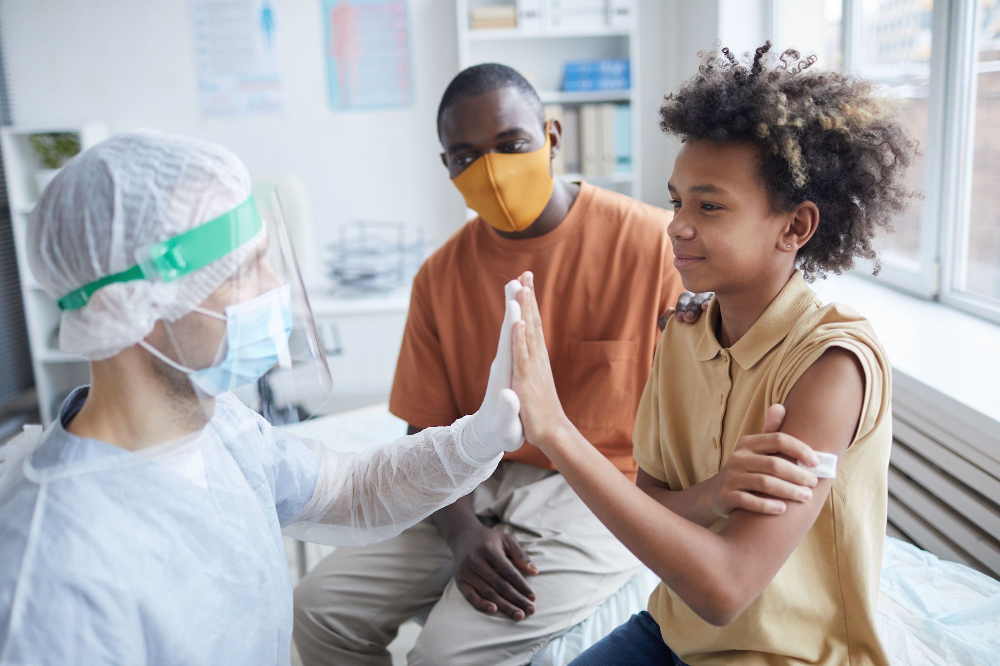A pediatric patient high-fives their healthcare provider, who is in full PPE, while their parent sits next to them.